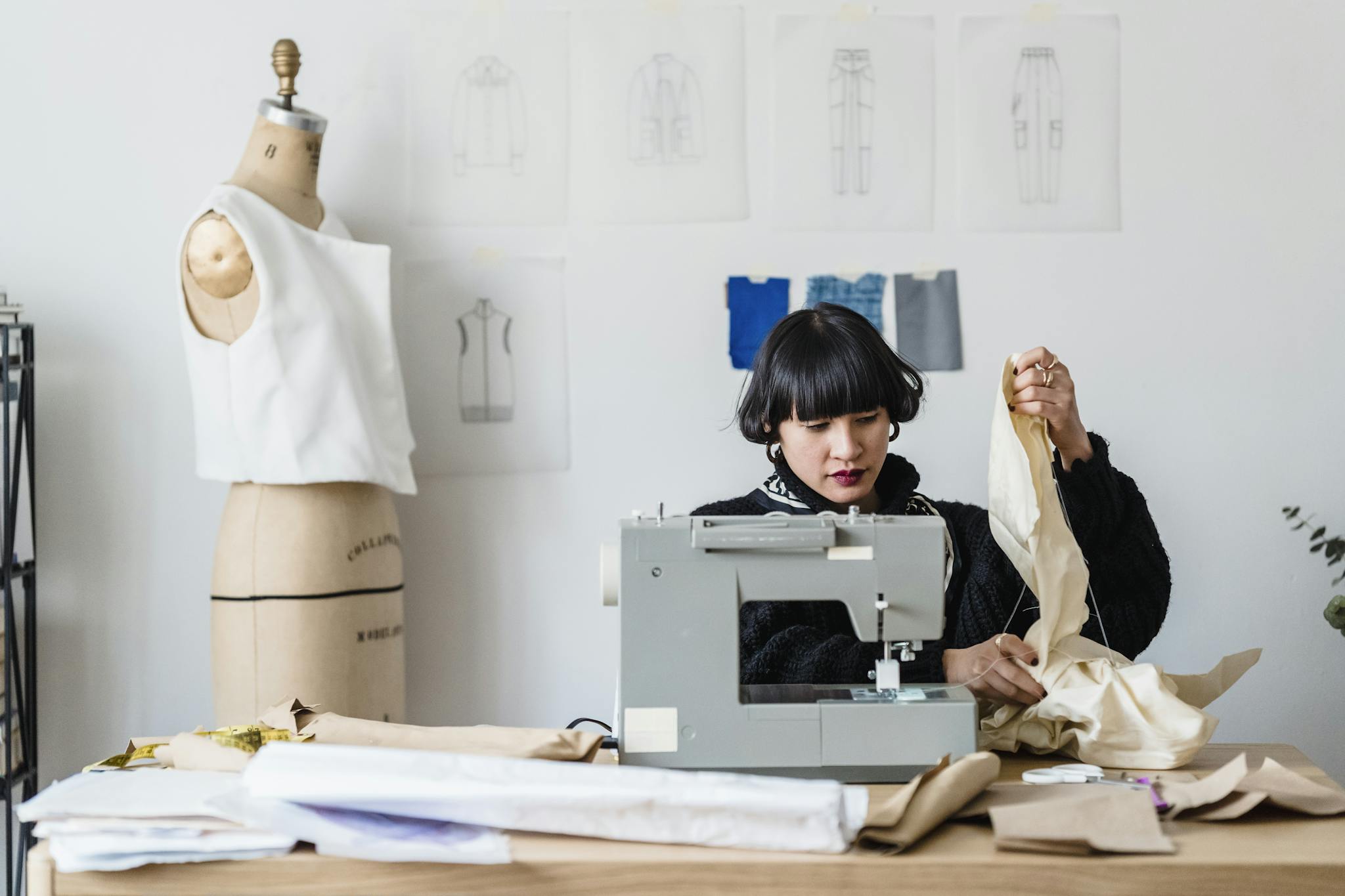 Concentrated fashionable young ethnic female designer with dark hair and red lips in warm sweater sewing dress with machine sitting at table with various materials in daylight in workshop